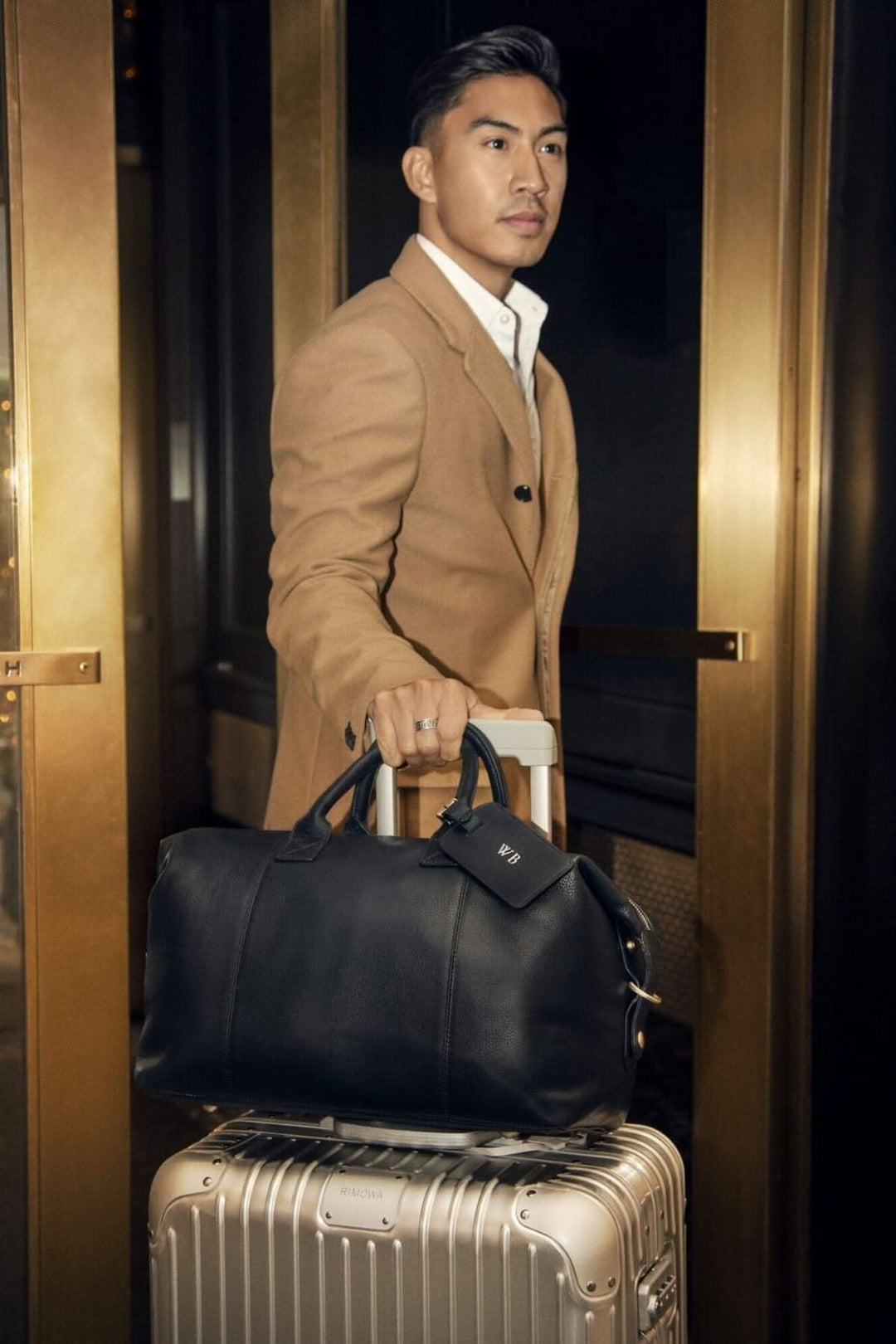 Homme élégant portant un sac de sport en cuir noir et tirant une valise dans un cadre lumineux.