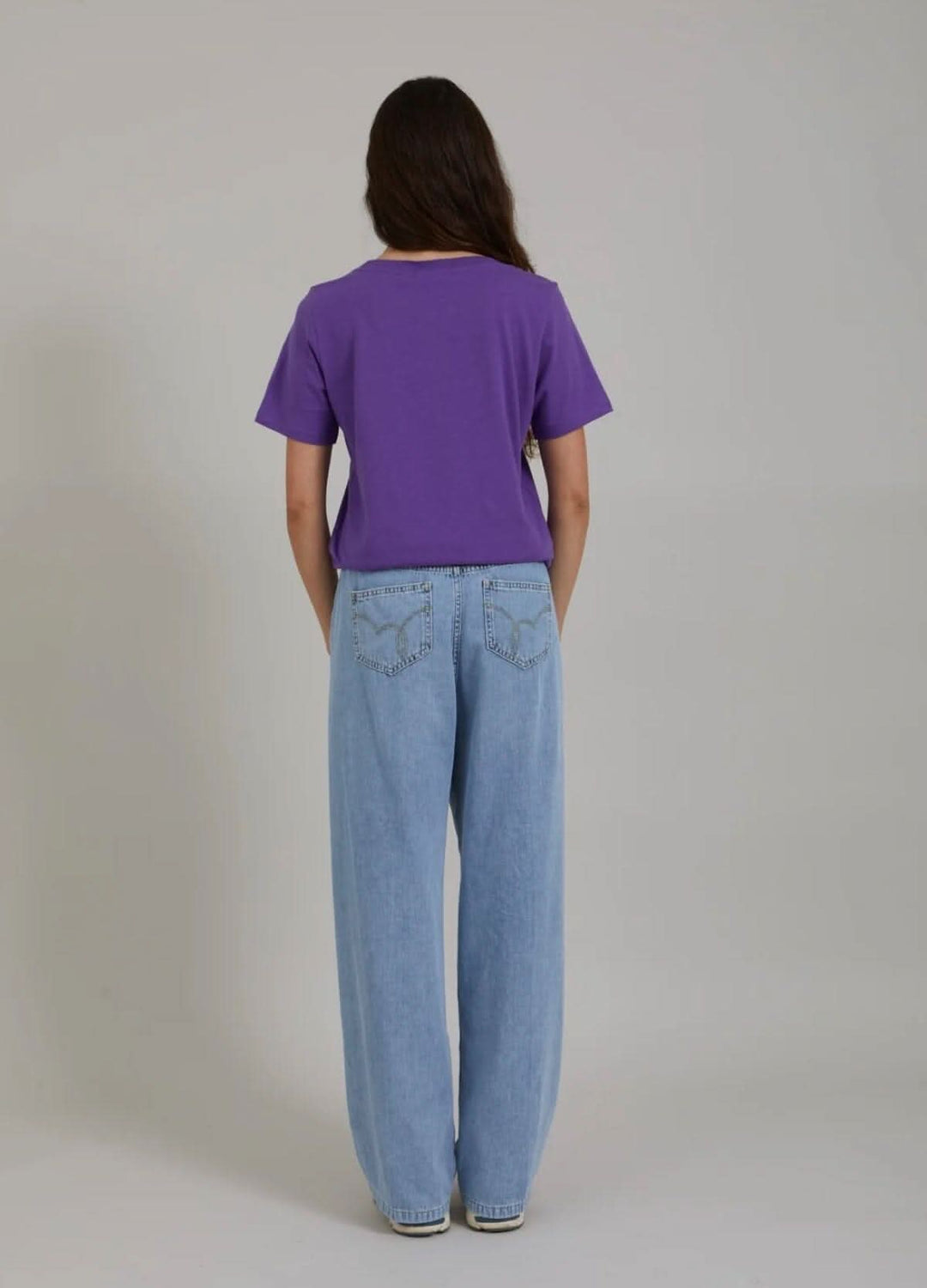 Femme vue de dos portant un t-shirt violet avec un pantalon bleu clair décontracté.