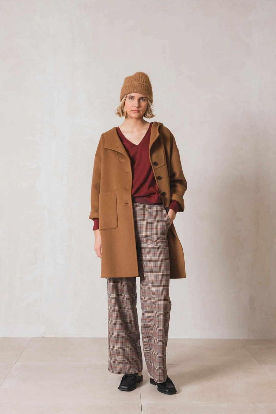 Femme portant un manteau marron avec un pull bordeaux et un pantalon à carreaux, style décontracté.
