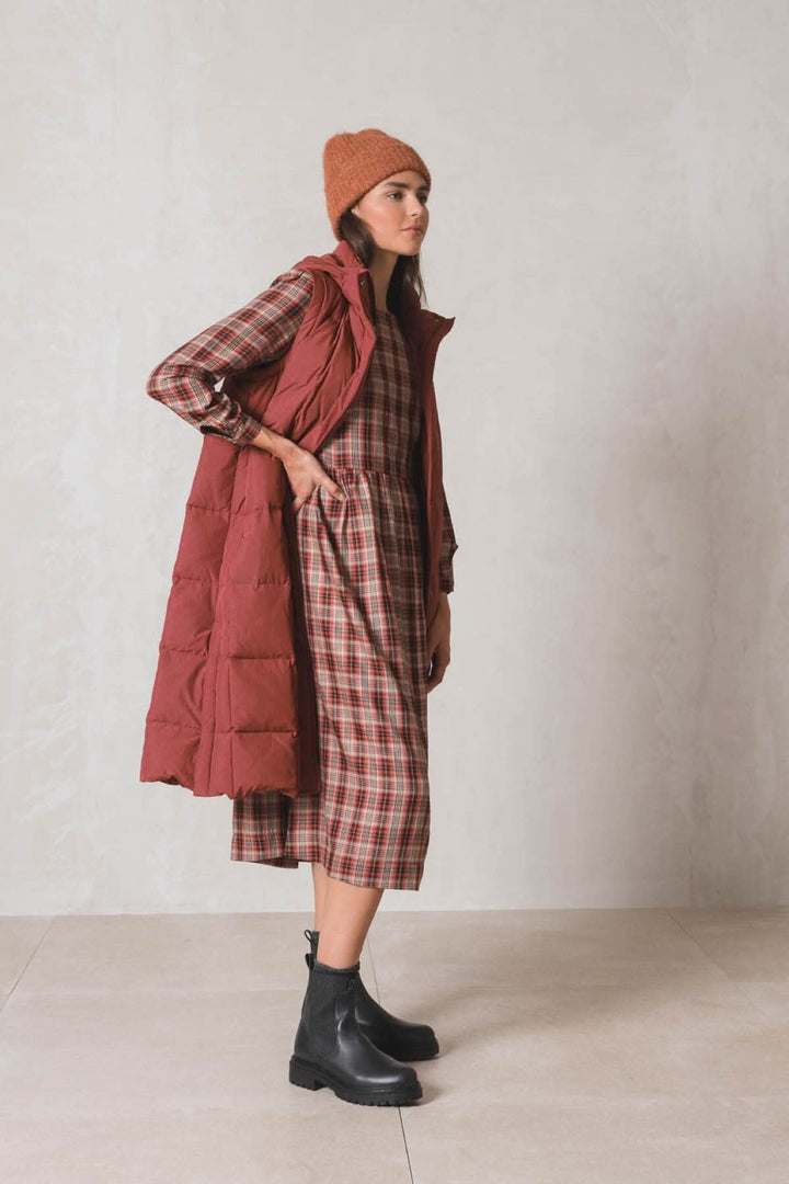 Femme portant une tuque en tricot et un gilet matelassé rouge avec une robe à carreaux, look automnal.