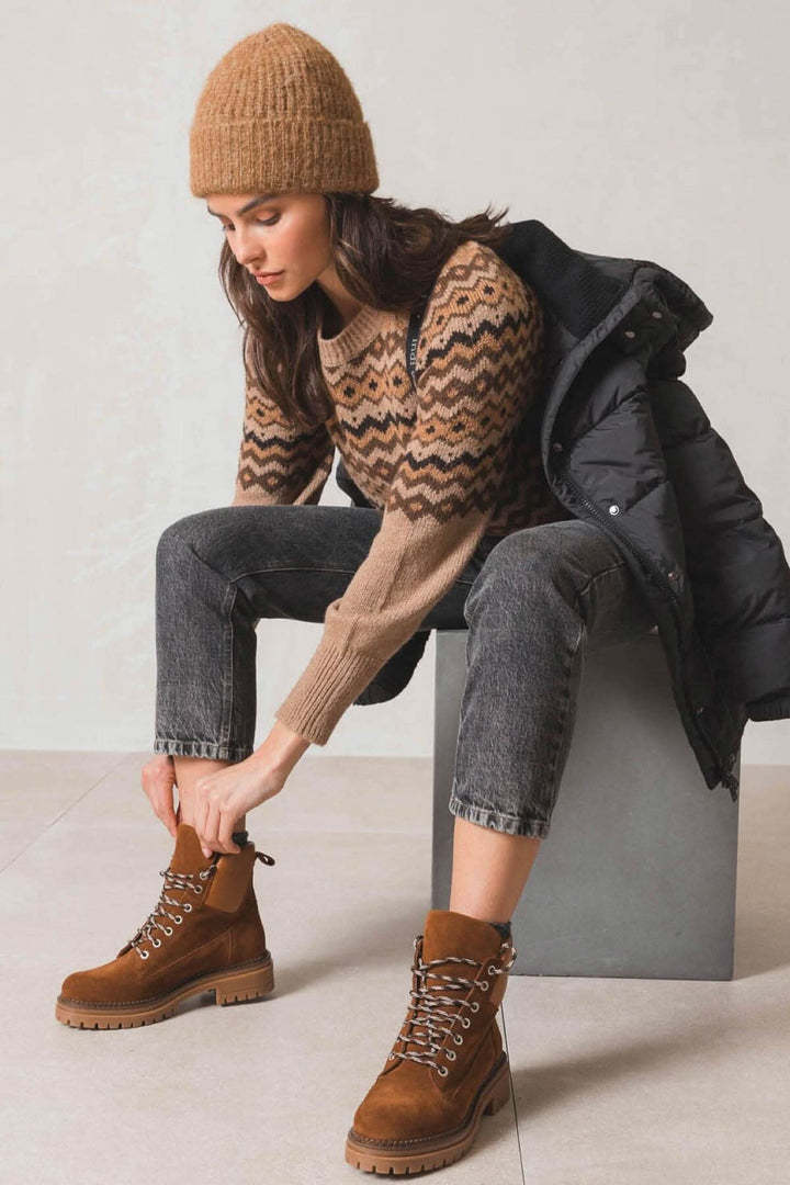 Femme stylée portant un pull en tricot, bottes marron et tuque, s'asseyant sur un cube en pierre.